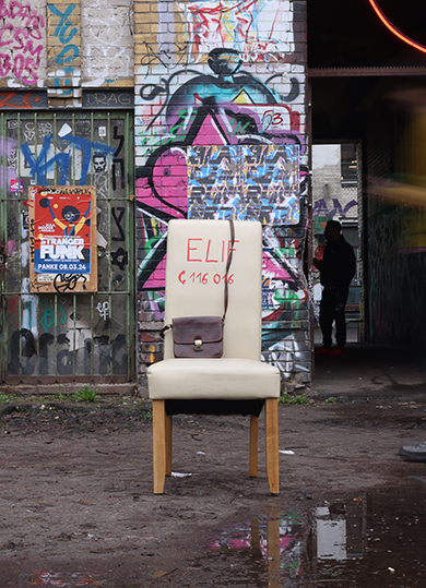 Vor einer Wand mit viel Graffiti steht ein weißer Stuhl mit Aufschrift Elif und der Notrufnummer 115016. Ein braune Handtasche liegt darauf.