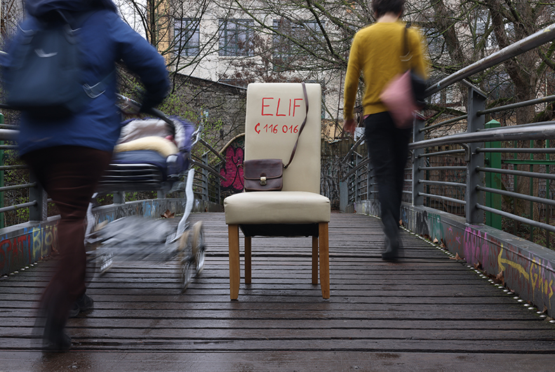 Auf einer Fußgängerbrücke im Park steht ein weißer Stuhl mit Aufschrift Elif und der Notfallnummer 116 016. Menschen gehen achtlos vorbei.