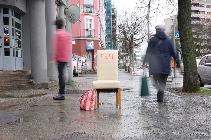 Auf einem Gehweg steht ein weißer Stuhl  mit Aufschrift Feli. Menschen gehen achtlos vorbei.