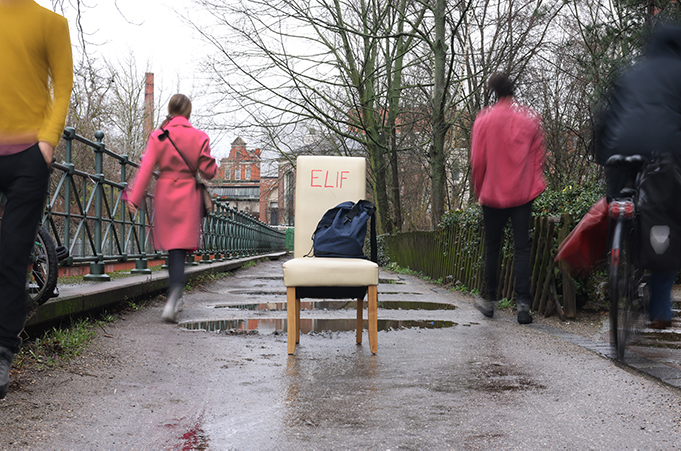 Auf einem nassen Gehweg steht ein weißer Stuhl mit Aufschrift Elif und einem blauen Rucksack. Mehrere Menschen gehen achtlos vorbei.