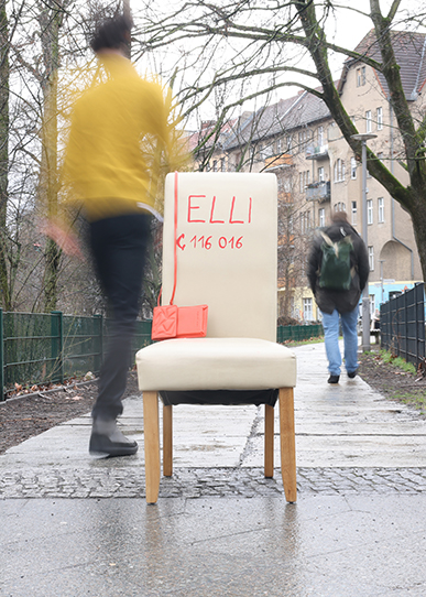 Auf einem Parkweg steht ein weißer Stuhl mit Aufschrift Elli und der Nummer des Hilfetelefons 116016 und einer roten Handtasche. Mehrere Menschen gehen achtlos vorbei.