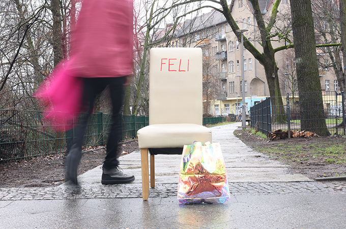 Auf einem Parkweg steht ein weißer Stuhl mit Aufschrift Feli, ein Einkaufsbeutel steht davor. Eine rosagekleidete Person geht daran vorbei.