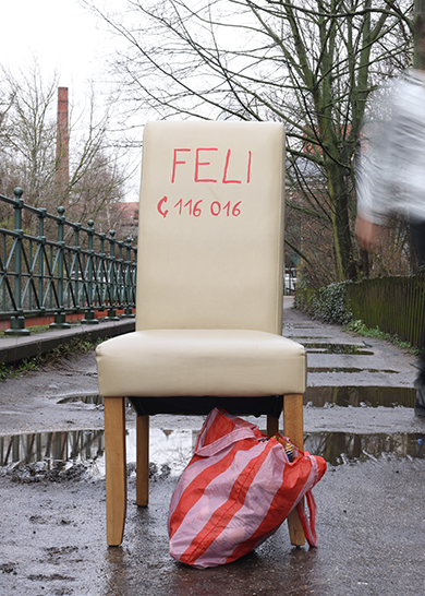 Auf einem Parkweg mit Pfützen steht ein weißer Stuhl mit Aufschrift Elif und der Notrufnummer 1160116. An den Stuhlbeinen lehnt eine rotgestreifte Tasche.