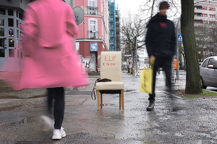 Auf einer Straße steht ein weißer Stuhl mit der Aufschrift Elif und der Nummer des Hilfetelefons 116016. Mehrere Leute laufen achtlos vorbei.