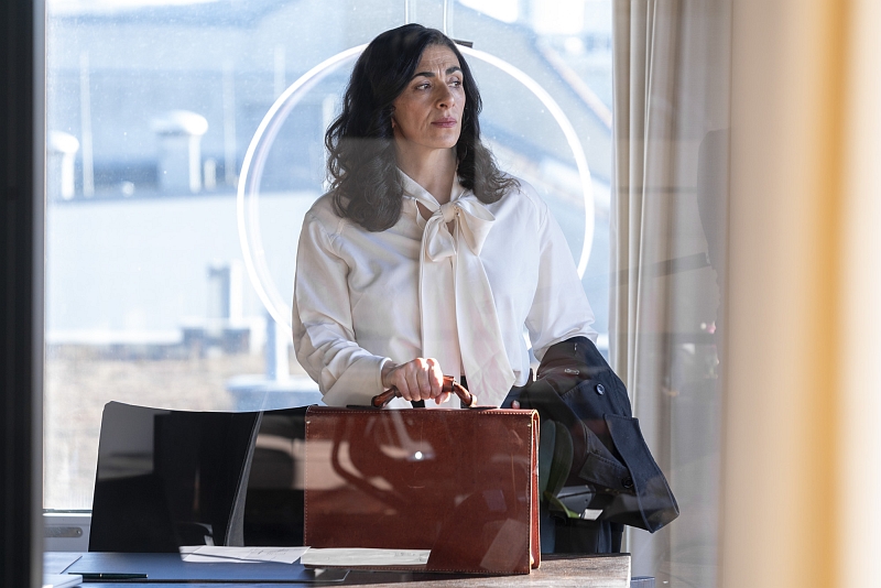 Dunkelhaarige Frau mit heller Bluse steht nachdenklich mit Aktentasche am Schreibtisch