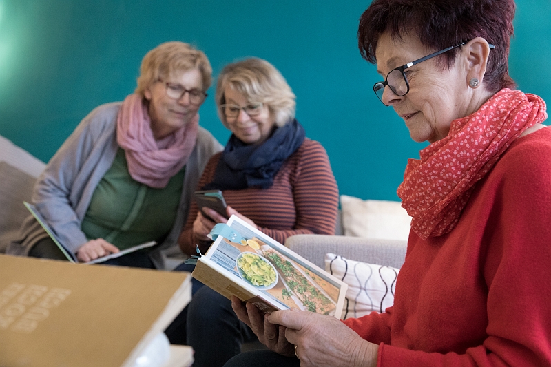 Drei alte Frauen suchen in Buch und Handy nach Kochrezepten