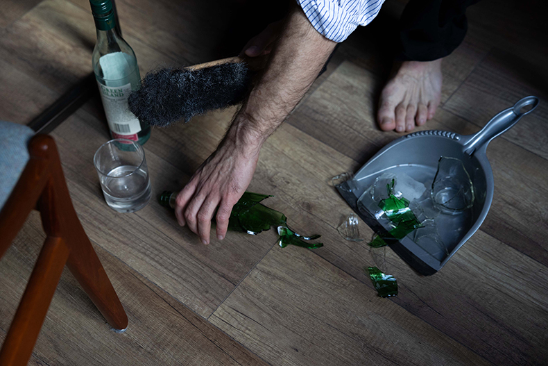 Männerhände legen Glasscherben in eine Kehrschaufel. Eine Schnapsflasche und ein Glas stehen daneben.