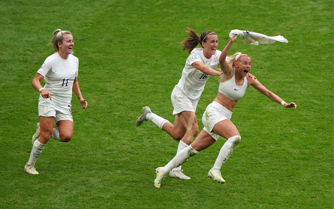 Die englische Fußballspielerin Chloe Kelly schwingt ihr Trikot über ihren Kopf. Sie rennt im Sport-BH übers Spielfeld, zwei weitere Teamkolleginnen voller Freude hinterher.