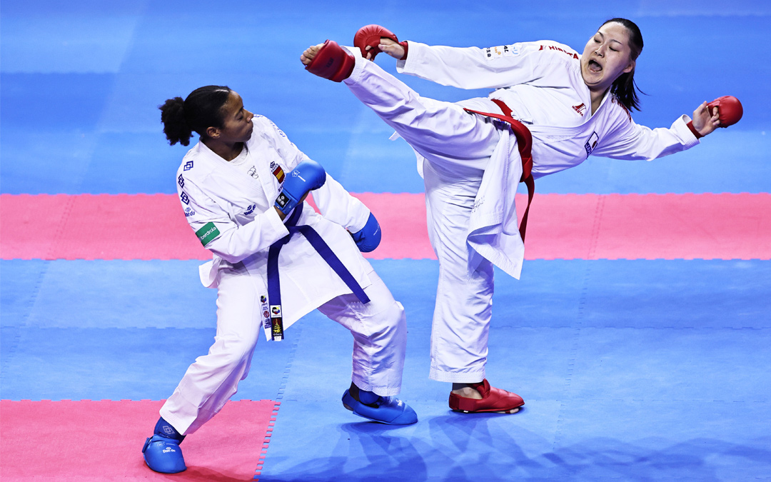 Eine Frau im weißen Karate-Anzug macht einen sportlichen Kumitetritt gegen den Oberkörper ihrer Gegnerin.