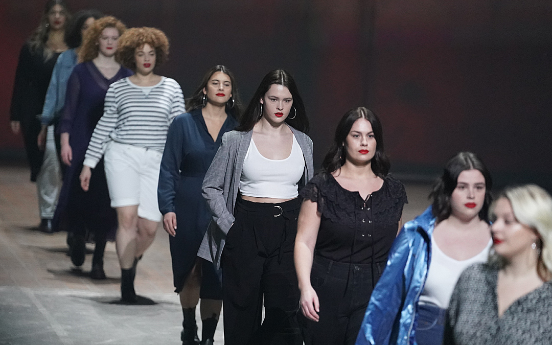 Bei einem Catwalk laufen neun junge Frauen in einer Reihe. Sie präsentieren modische Alltagskleidung. Keine hat die üblichen dünnen Modelmaße