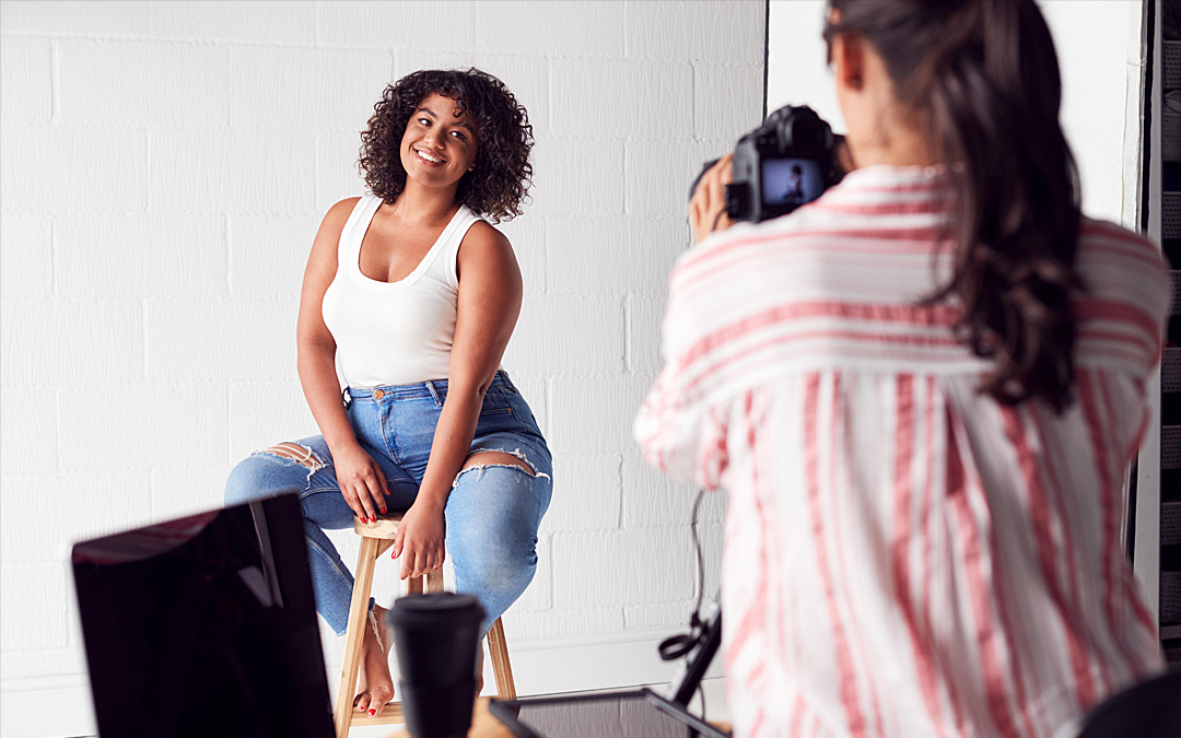 Junge Frau mit weißem Tanktop und in Jeans sitzt im Fotostudio auf einem Hocker. Die Fotografin in Rückenansicht