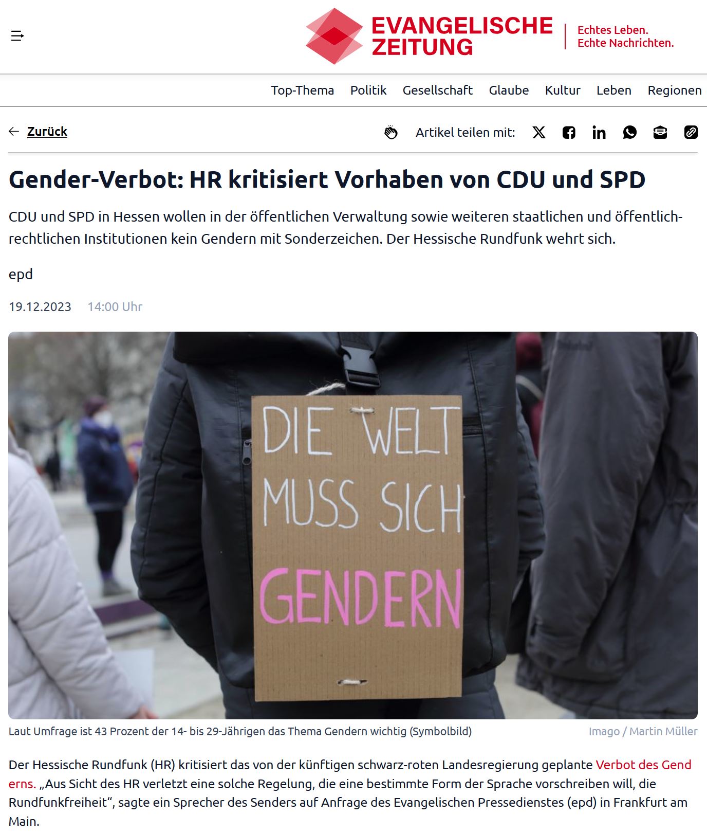 Screenshot mit Foto von einer Demo: Eine Person in schwarzer Winterjacke hat sich ein Schild umgehängt mit Text: Die Welt muss sich gendern.