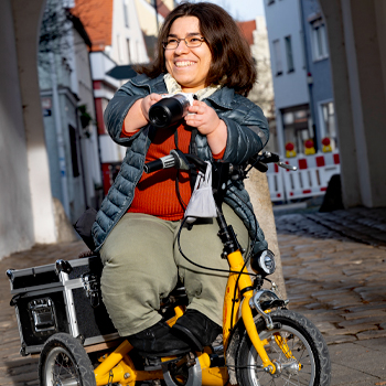 Kleinwüchsige Frau mit dunklen Haaren und Brille sitzt auf ihrem Laufrad und hält eine Fotokamera in den Händen.