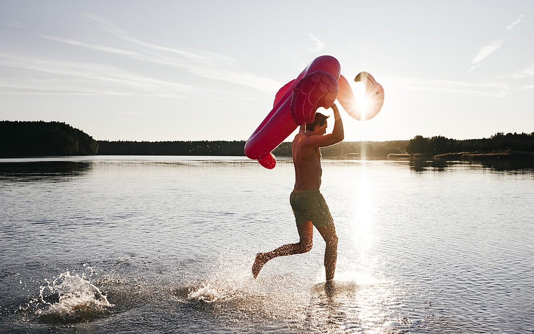 Ein Mann rennt mit einem Flamingo-Schwimmreifen durchs Flachwasser eines Sees. Die Sonne strahlt.