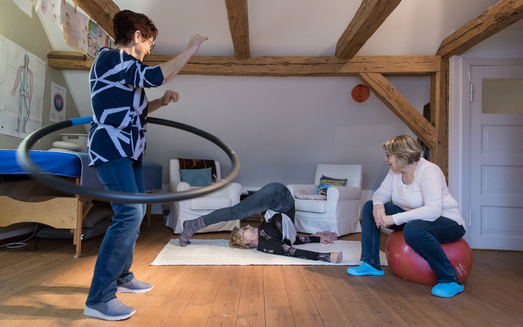 Gymnastik im Wohnzimmer: Eine alte Frau mit einem Hula-Hup-Reifen, eine macht eine Brücke auf dem Boden, die dritte guckt zu auf einem Sitzball