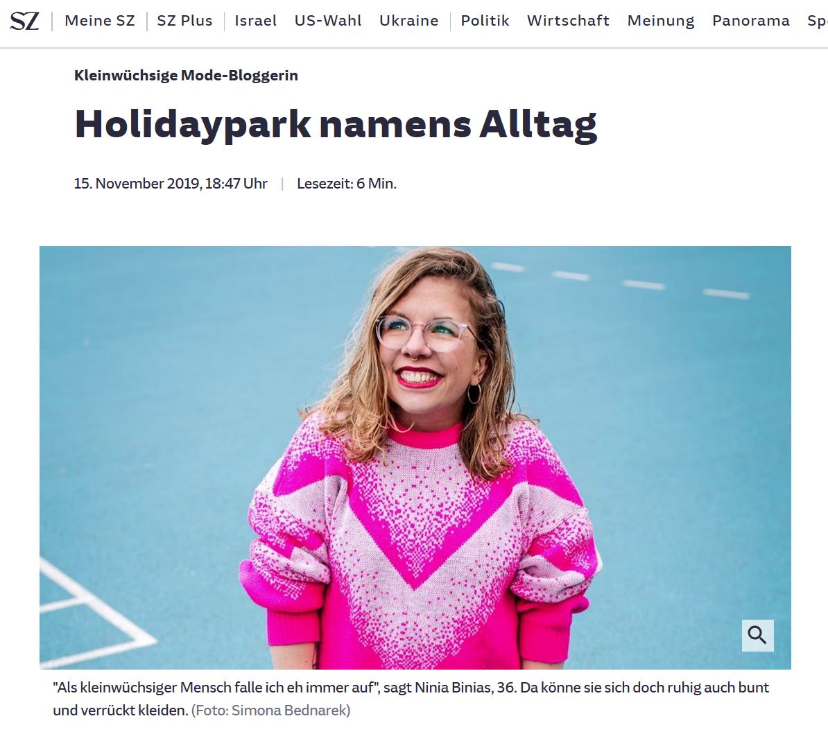 Screenshot mit Portraitfoto von Ninia LaGrande. Sie hat lange blonde Haare, Brille und trägt roten Lippenstift zu einem rot-pinken Pullover