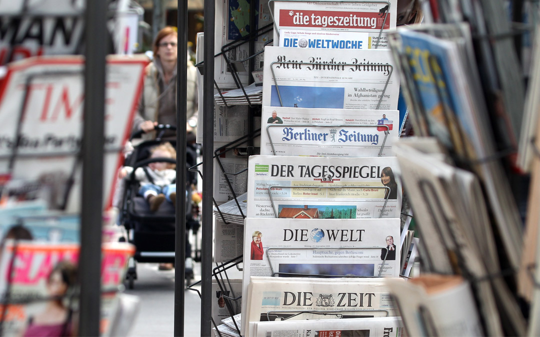 Zeitungsständer an einem Kiosk, dazwischen erkennbar läuft eine Frau mit einem Kinderwagen